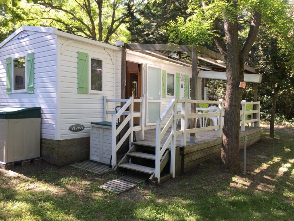 Mobil-home IRM blanc, volets verts et terrasse bois avec balustrade blanche sous les arbres en camping.